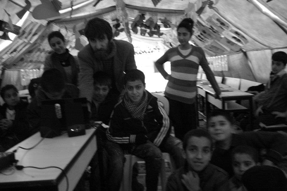 Siyah beyaz fotoğrafta Samet Aksuoğlu hafif kambur durarak deprem mağduru çocuklarla birlikte film seyrediyor. Fotoğraf sanki perdenin içinden çekilmiş gibi herkes kadraja bakıyor.